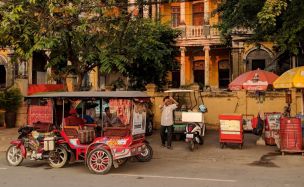 Vor einem Haus in Kambodscha sind kleine Verkaufsstände aufgebaut, ein Motorrad mit Passagierkabine steht bereit.
